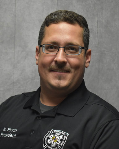 A man with short dark hair, glasses, and a mustache wears a black collared shirt with an embroidered badge and "N. Ervin President" on it, posing in front of a plain gray background.