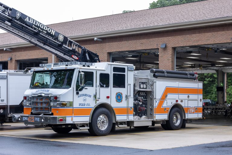 A white and orange fire truck labeled “Abingdon Fire Co.” is parked outside a brick fire station with open garage bays. The truck features ladders and hoses, and part of a ladder truck is visible behind it.