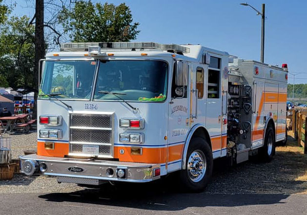 A white and orange fire truck is parked outdoors on a sunny day, with trees and picnic tables visible in the background. The fire truck has emergency lights and various equipment compartments.
