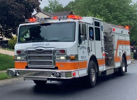 A white fire truck with orange stripes and flashing lights is parked on a residential street on a cloudy day, with trees and houses in the background.