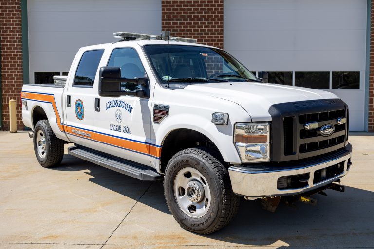 A white Ford pickup truck with blue and orange stripes and “Hyndman Fire Co.” insignia is parked in front of a building with white garage doors and brick walls.