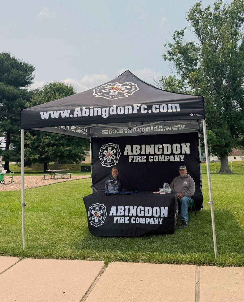 Two people sit at a table under a black canopy tent with "Abingdon Fire Company" and its logo. The tent is set up on grass in a park-like area with trees and a playground in the background.