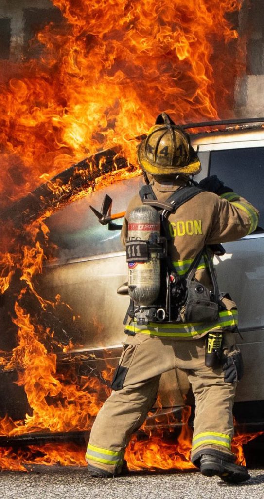 A firefighter in full gear stands near a burning car, facing intense orange flames and thick smoke, preparing to respond to the emergency.