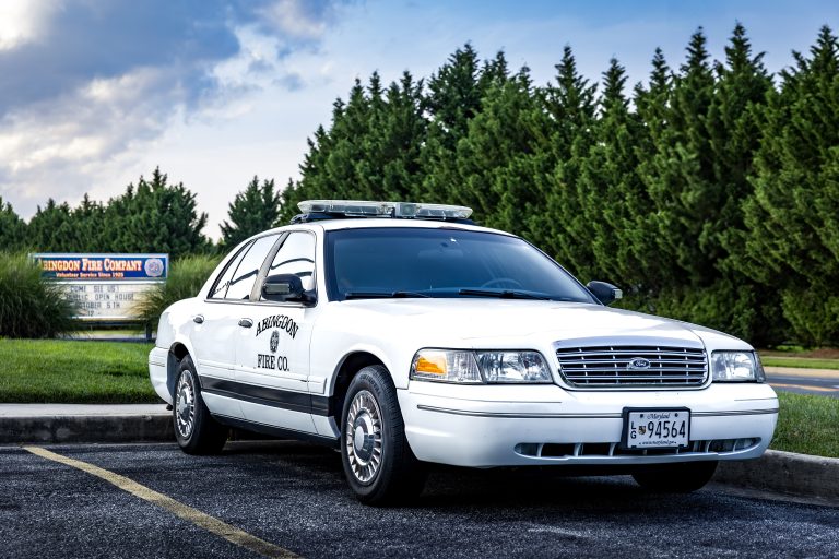 A white sedan marked "Fire Co." with emergency lights on the roof is parked in a lot near a sign for Rehoboth Fire Company, surrounded by green trees and under a partly cloudy sky.