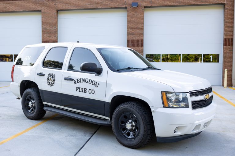 A white Chevrolet SUV with "Abingdon Fire Co." and a fire department emblem on the side is parked in front of a building with large garage doors.