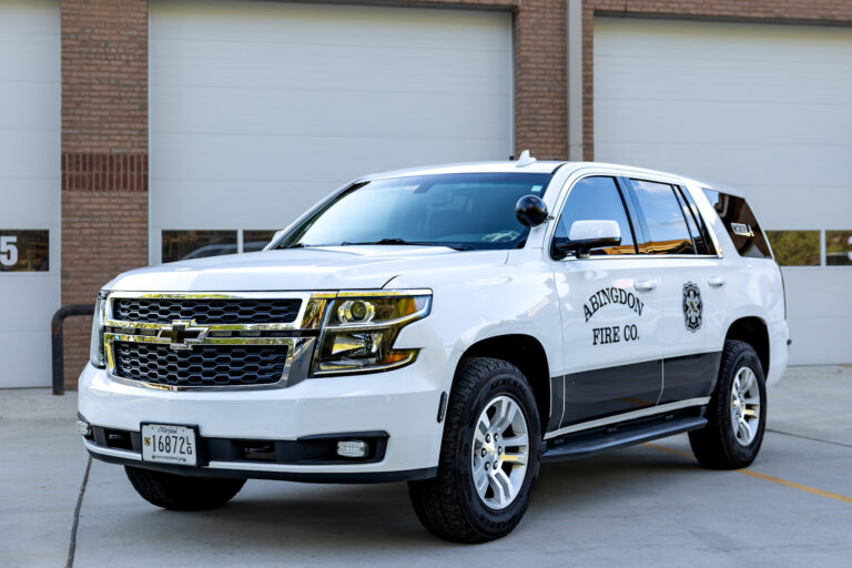 A white Chevrolet SUV labeled "Abingdon Fire Co." is parked outside a building with large garage doors. The vehicle has official markings and a Maryland license plate.