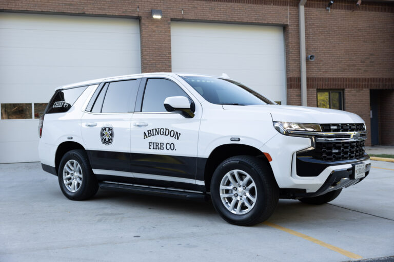 A white Abingdon Fire Company SUV is parked outside a brick building with large garage doors. The vehicle features the fire company emblem and text on the side.