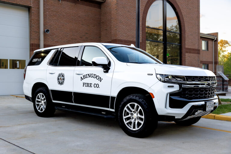 A white and black SUV labeled "Abingdon Fire Co." is parked outside a brick building, possibly a fire station. The vehicle has a fire department emblem on the door and emergency lights on the roof.