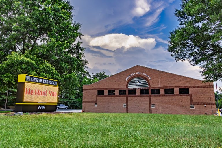 A brick fire station with a sign reading "We Want You" on the lawn. The building is labeled "Abingdon Fire Company" and is surrounded by green grass and trees under a partly cloudy sky.