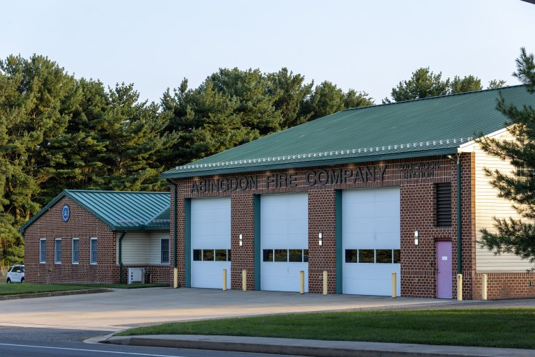 A brick fire station with a green metal roof, reading "Abingdon Fire Company," features four large white garage doors and is surrounded by trees on a clear day.