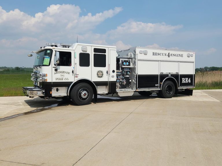 A white and black fire engine labeled "Rescue 4 Engine" and "RA4" is parked on a paved surface with grass and a clear blue sky in the background.