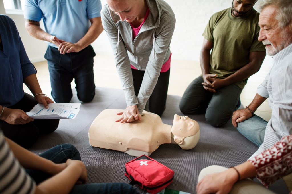 A group of people kneel around a CPR training manikin while one person demonstrates chest compressions. A first aid kit and instructions are visible on the floor.