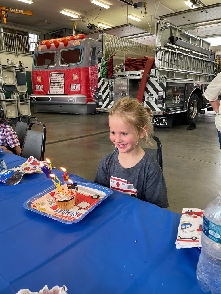 A young girl smiles at a cupcake with lit candles on a table, celebrating in a fire station with a fire truck in the background.