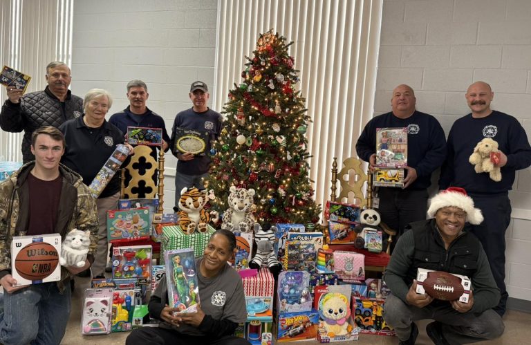 A group of people in matching shirts pose around a decorated Christmas tree surrounded by toys and gifts. They are smiling and holding toys, with wrapped presents and stuffed animals arranged in front.