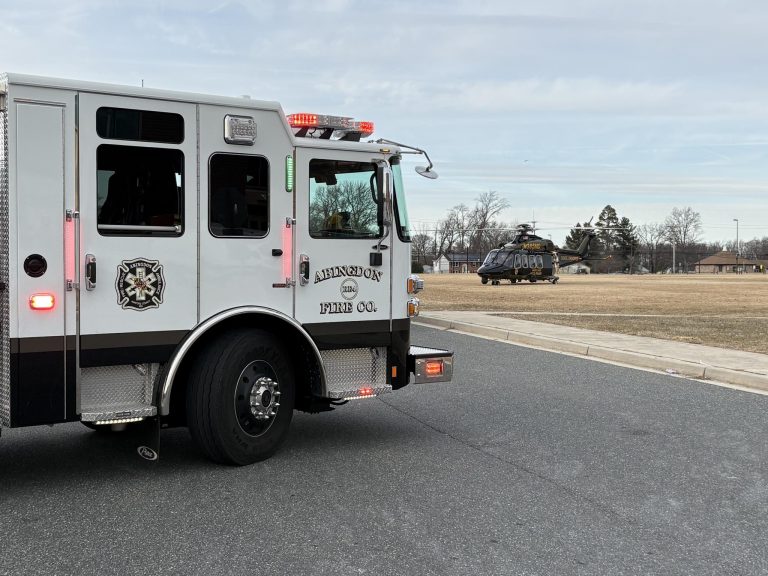 A Lindenwold Fire Co. fire truck is parked on a road near a grassy field where a helicopter is landed. The sky is cloudy and trees and buildings are visible in the background.