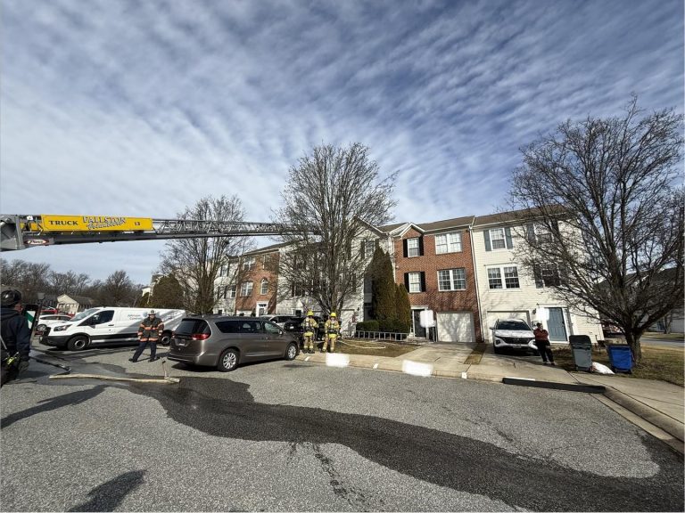 Firefighters stand outside a residential townhouse building as a fire truck ladder extends toward the roof. Several emergency personnel, vehicles, and a black van are visible in the parking area.