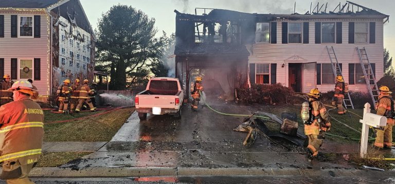 Firefighters extinguish a house fire at dusk, with smoke rising from a burned two-story home. Firefighters, hoses, and a white pickup truck are visible in the driveway; neighboring houses and trees are in the background.