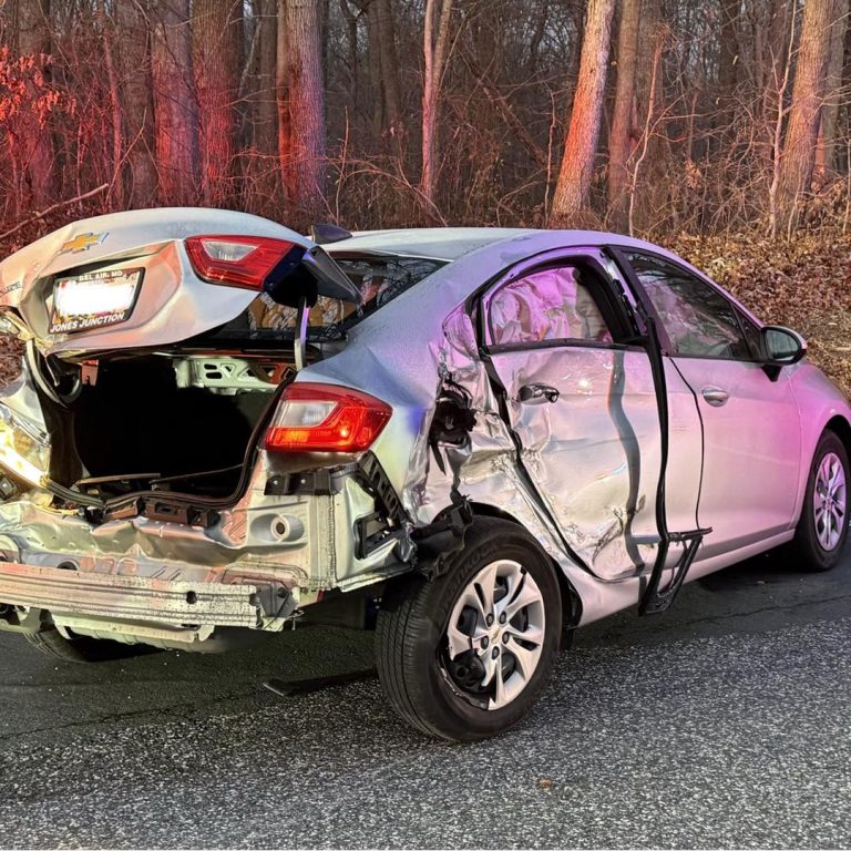 A silver car with severe rear and side damage is parked on a road near a wooded area. The trunk is crumpled, the rear bumper is hanging, and the left side doors are badly dented.