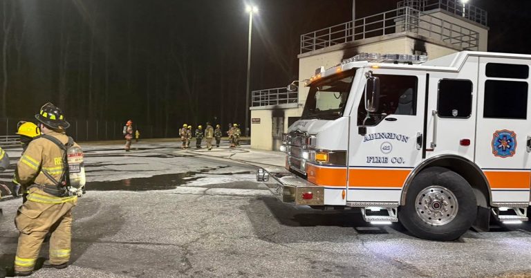 Several firefighters in gear stand and walk near a white and orange fire truck marked “Aberdeen Fire Co.” at night in a well-lit parking lot, with a building in the background.