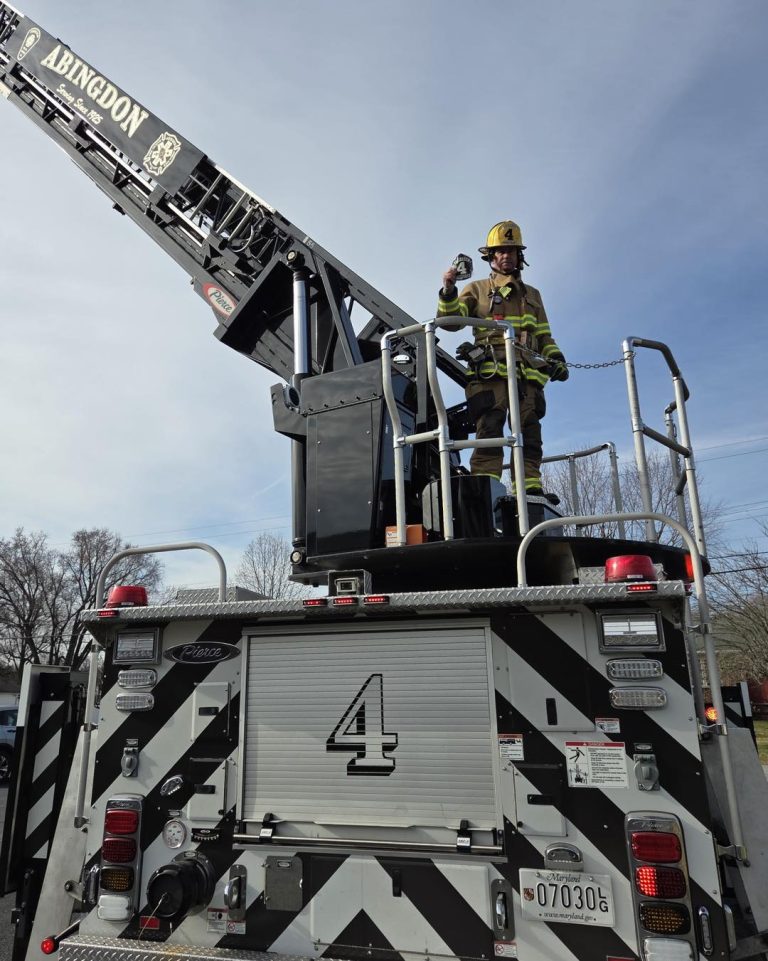 A firefighter in full gear stands on the raised platform of a fire truck with “Abingdon” on the ladder. The firefighter is holding a drink and waving. The back of the truck displays the number 4. Trees and blue sky are visible.