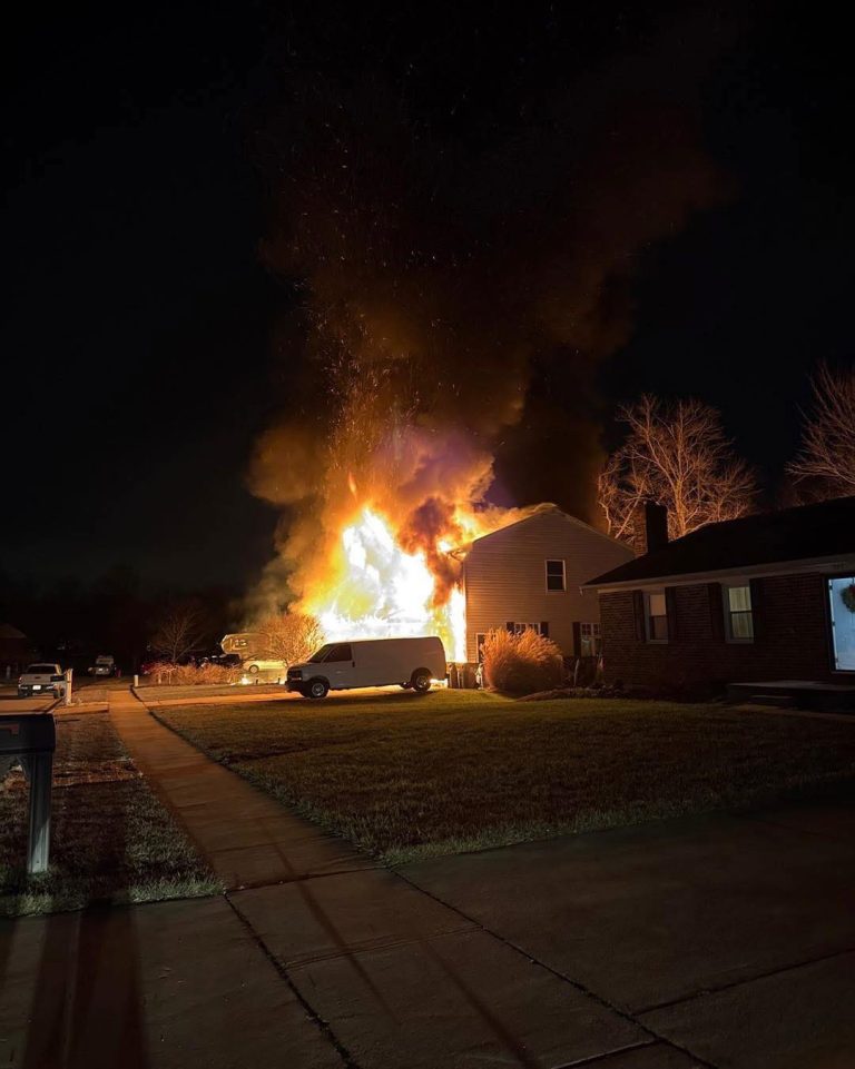 A large fire engulfs the side of a house at night, with tall flames and thick smoke rising into the sky. A white van is parked in the driveway, and nearby homes appear untouched by the blaze.