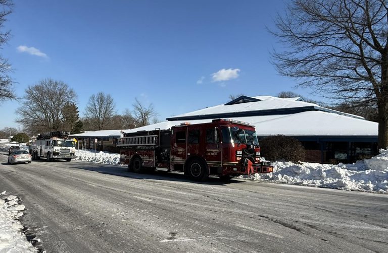 A red fire truck and a white utility truck are parked on a snowy street in front of a building with a snow-covered roof under a clear blue sky. Snow piles line the road, and bare trees stand nearby.