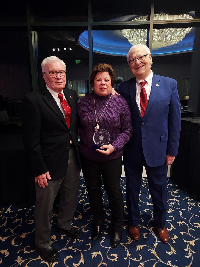 Three people stand together indoors, smiling. The woman in the center holds a round glass award. The two men on either side wear suits and ties. They are standing on a patterned carpet in front of large windows.