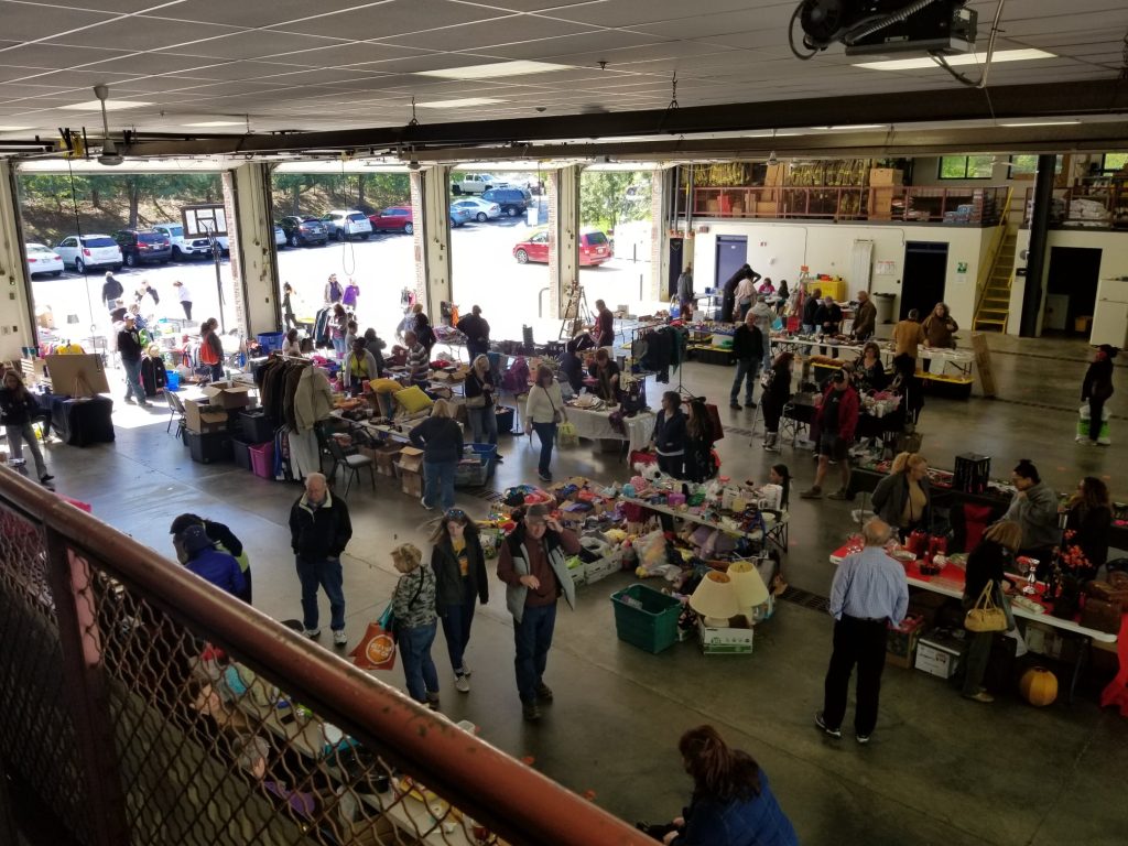 A community indoor flea market with people browsing tables filled with various items in a large open garage space. Sunlight streams through open doors, and cars are parked outside.