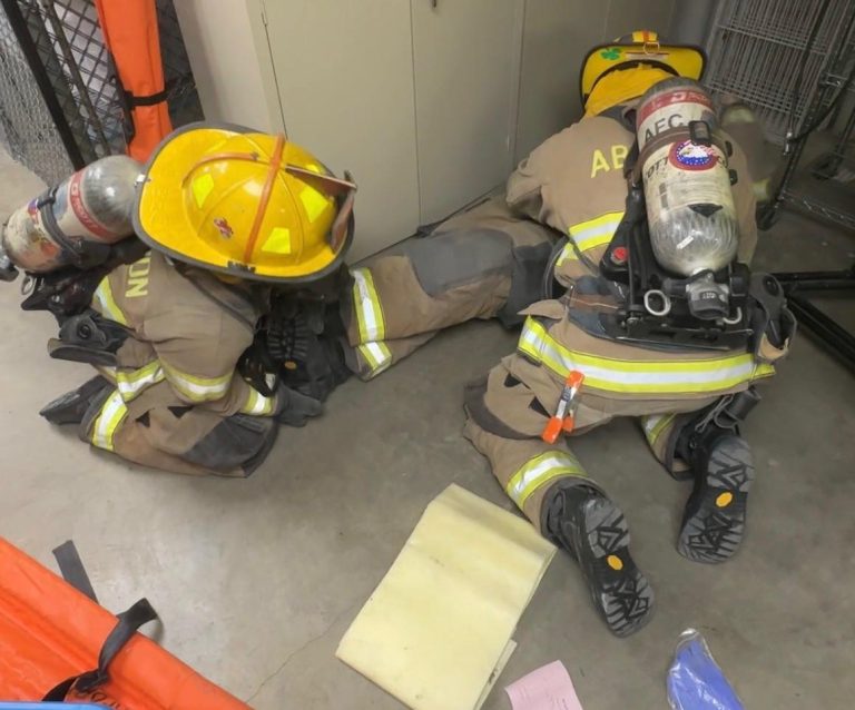 Two firefighters in full gear and helmets are crouched on the ground indoors, appearing to assist each other. Papers and protective padding are scattered nearby on the concrete floor.