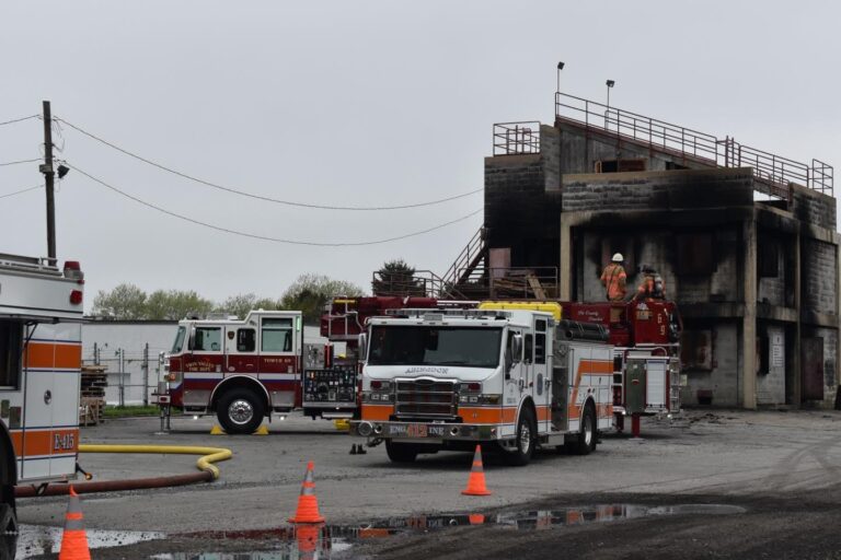 Three fire trucks are parked near a concrete training building with burn marks. Firefighters in gear are on a truck’s ladder, appearing to conduct a drill. Orange traffic cones are set up around the scene on a cloudy day.