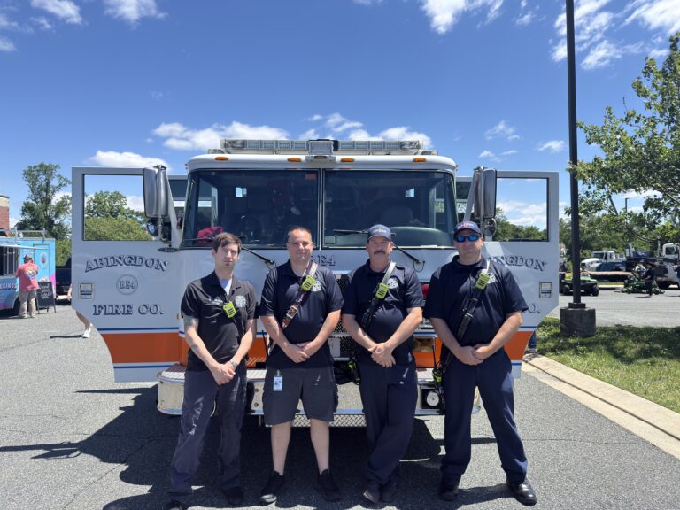 Four firefighters stand in front of a fire truck labeled "Abingdon Fire Co." on a sunny day, posing together in uniform in a parking lot with trees and other vehicles in the background.