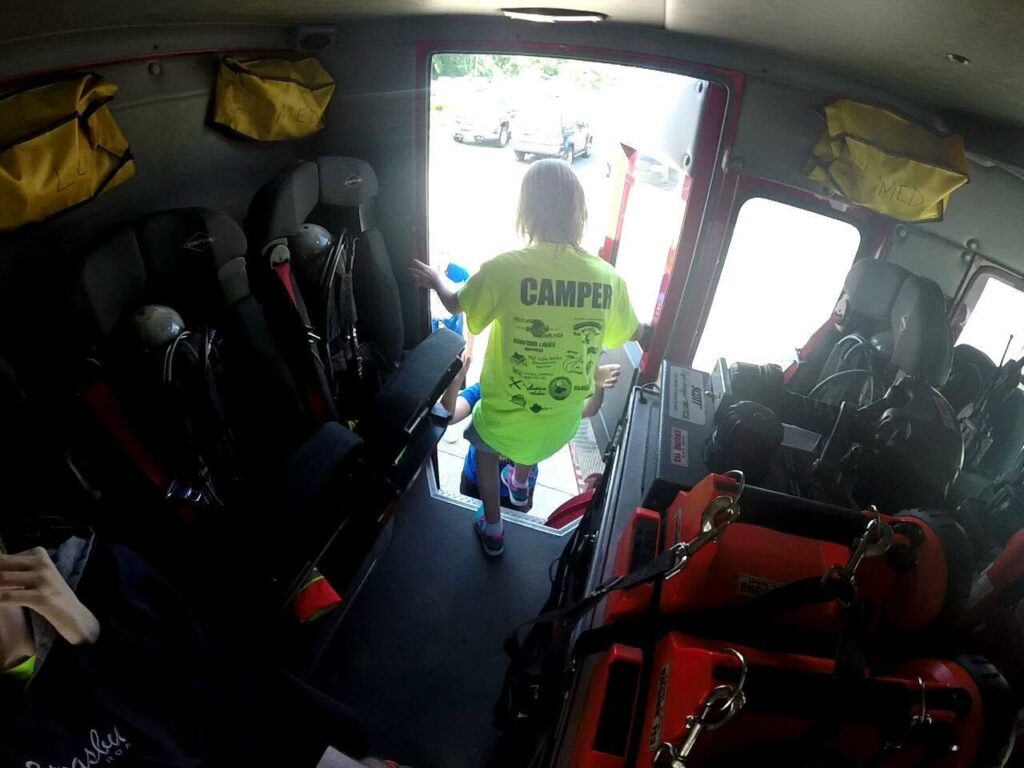 A child wearing a bright yellow "CAMPER" shirt stands at the open door of a vehicle, possibly a fire truck, with various gear and seats visible inside. Sunlight shines outside where cars are parked.