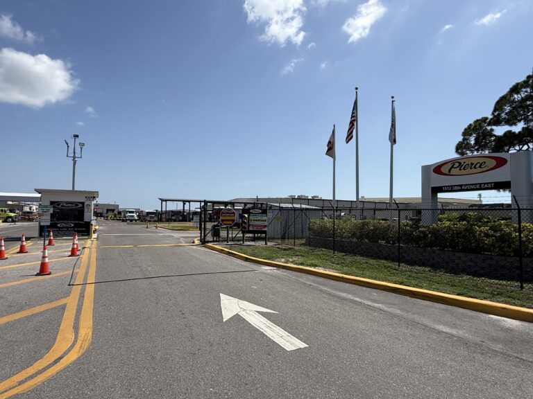 Entrance to an industrial facility with traffic cones, security gate, three flags, and a sign reading "Pierce." Blue sky with clouds is visible above.