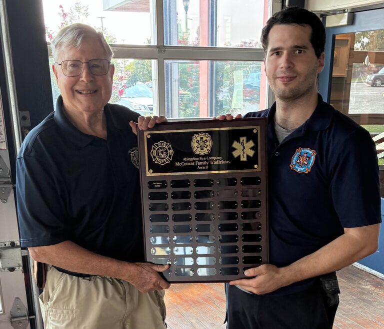 Two men in dark uniforms stand indoors, smiling and holding a commemorative plaque with fire and medical symbols. Sunlight shines through large windows behind them, and trees are visible outside.