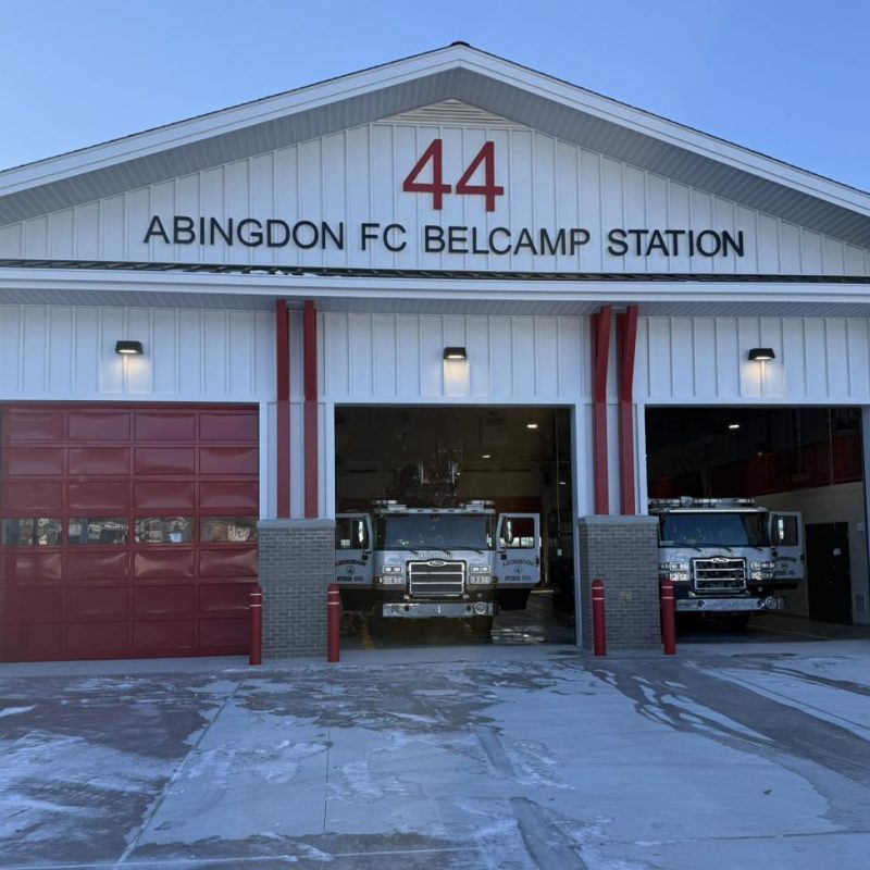 A fire station labeled "44 ABINGDON FC BELCAMP STATION" with three garage doors, two of which are open showing fire trucks inside. The station has a white exterior with red accents.