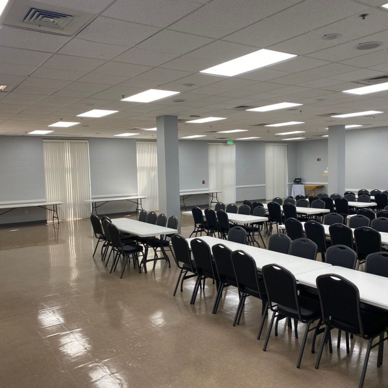 A spacious, well-lit room with several rows of long white tables and black chairs arranged for a meeting or event. The floor is shiny, and the walls are light-colored with large windows and ceiling lights.