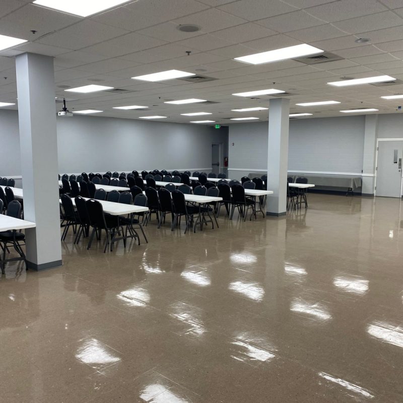 A large, empty room with rows of white tables and black chairs arranged on the left side. The floor is shiny and brown, and the ceiling has fluorescent lights. Doors and a trash can are visible on the right wall.