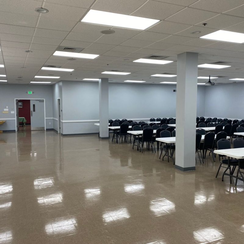 A large, brightly lit room with rows of empty black chairs and white rectangular tables on one side. The floor is shiny, and the walls are light-colored. There's a small table near a door on the left.