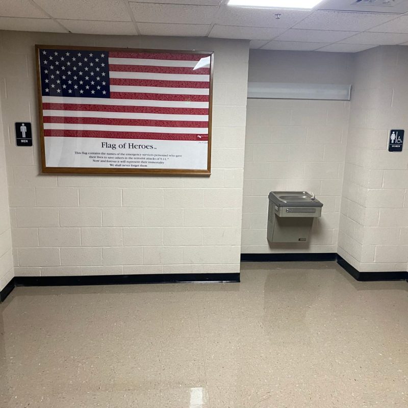 A hallway with red doors to men's and women's restrooms, a drinking fountain, and a large framed American flag labeled "Flag of Heroes" on the wall. The floor is shiny and the walls are light-colored.