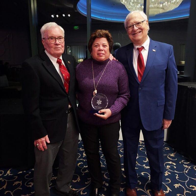 Three people stand together indoors, smiling. The woman in the center holds a round glass award. The two men on either side wear suits and ties. They are standing on a patterned carpet in front of large windows.