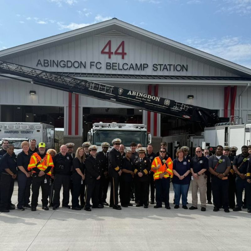 A large group of firefighters and emergency personnel stand in front of the Abingdon FC Belcamp Station 44, with fire trucks and equipment visible behind them on a sunny day.