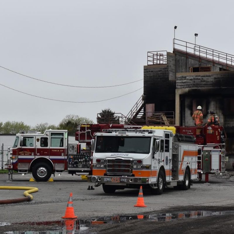 Three fire trucks are parked near a concrete training building with burn marks. Firefighters in gear are on a truck’s ladder, appearing to conduct a drill. Orange traffic cones are set up around the scene on a cloudy day.