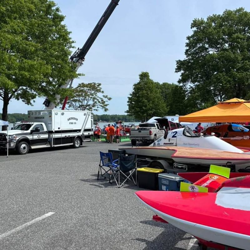 Several powerboats are parked on trailers in a lot near a lake, with an orange canopy, folding chairs, and a fire department crane truck visible. People gather in the background among green trees on a sunny day.