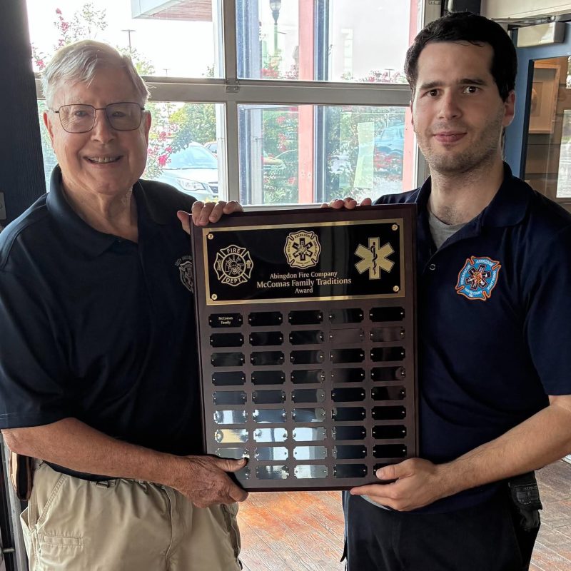 Two men in dark uniforms stand indoors, smiling and holding a commemorative plaque with fire and medical symbols. Sunlight shines through large windows behind them, and trees are visible outside.