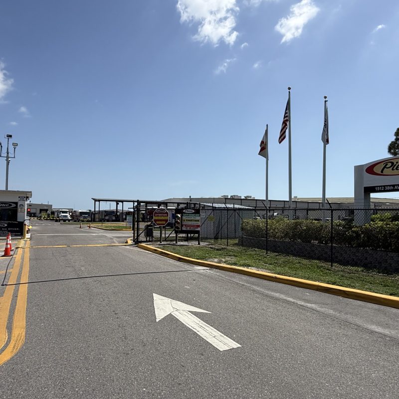 Entrance to an industrial facility with traffic cones, security gate, three flags, and a sign reading "Pierce." Blue sky with clouds is visible above.