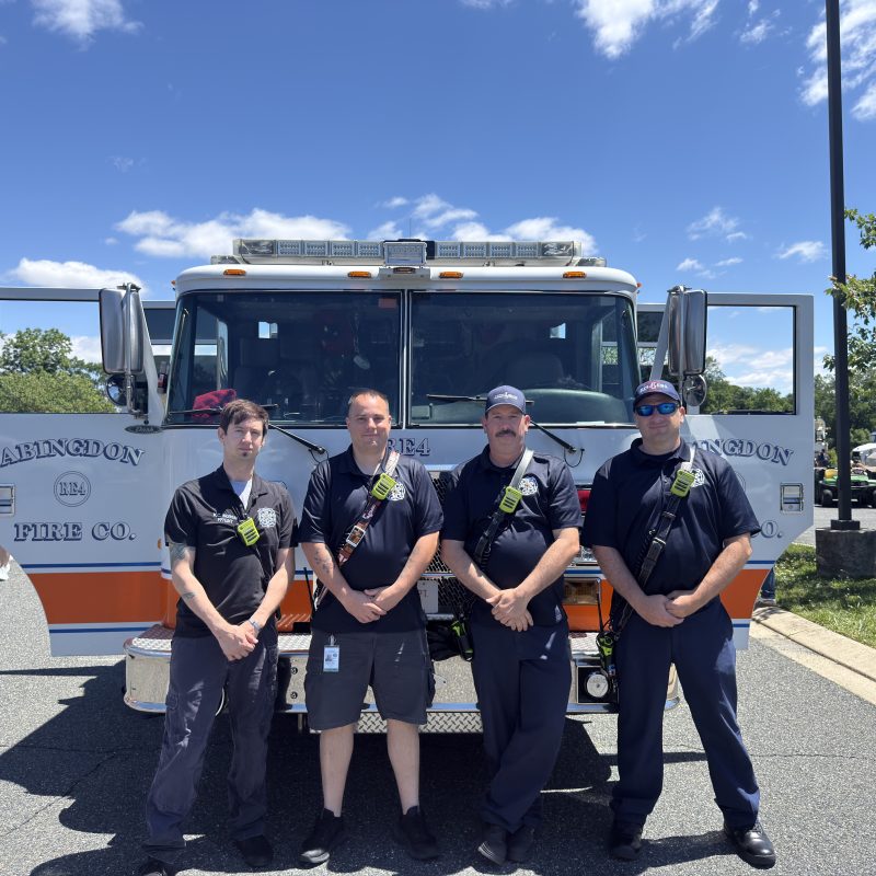 Four firefighters stand in front of a fire truck labeled "Abingdon Fire Co." on a sunny day, posing together in uniform in a parking lot with trees and other vehicles in the background.