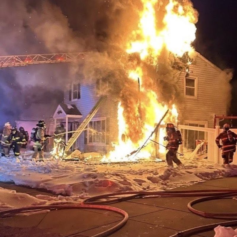 Firefighters work to extinguish a large blaze engulfing the side of a two-story house at night. Flames and smoke rise high, while hoses and snow cover the ground in the foreground.